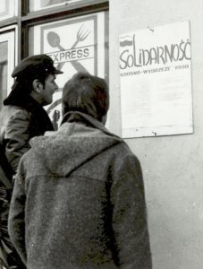 Zdjęcie dwóch mężczyzn, którzy czytają ogłoszenie zatytułowane Solidarność Krosno-Wybrzeże 1980 - wywieszone na ścianie budynku