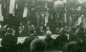 Fotografia uroczystości w fabryce z ok 1937 r. grupa osób siedząca przy stole, nad którym widać kopię balonu wyprodukowanego w Sanoku