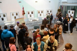 młodzież zwiedzająca wystawę, w tle gabloty z dokumentami archiwalnymi i portrety żołnierzy z okresu międzywojennego oraz manekin w mundurze 20 Pułku Ułanów