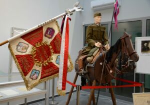 manekin w mundurze żołnierza z okresu miedzywojennego siedzący na atrapie konia i sztandar 20 pułku Ułanów